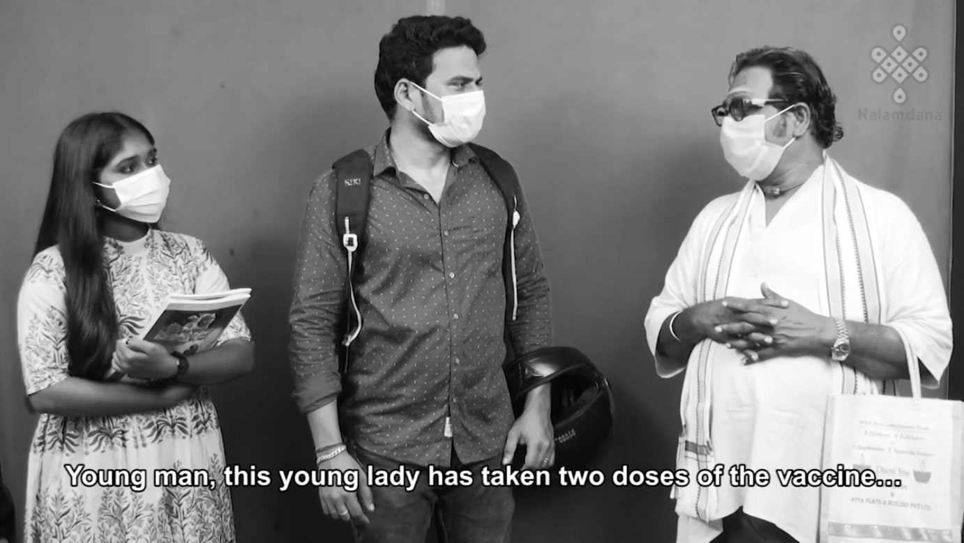 Three Tamil people wearing masks. The man on the right is saying "Young man, this young lady has taken two doses of the vaccine..."